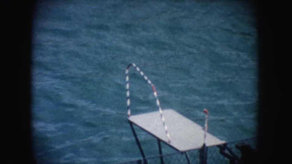 1962 California home-movie frame with a diving platform, striped handrail, and rippling blue water in the pool.