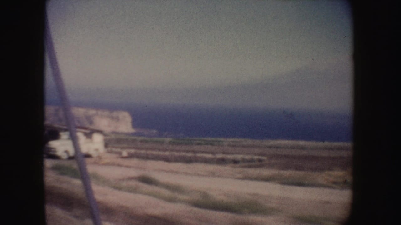 1962 California 8mm frame showing a dirt coastal road, blue ocean, pale cliffs, and a white vehicle near the left edge.