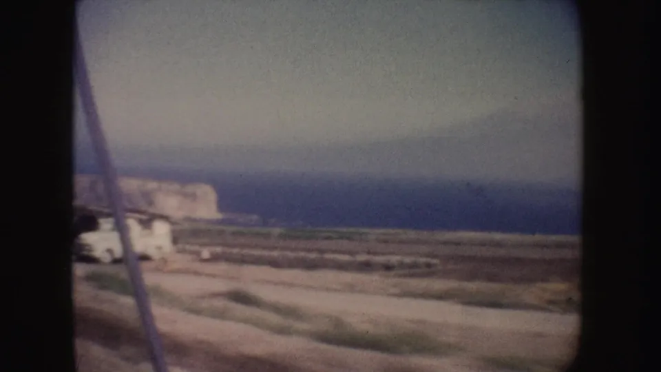 1962 California 8mm frame showing a dirt coastal road, blue ocean, pale cliffs, and a white vehicle near the left edge.