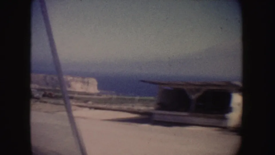 1962 California coastal roadside view with a parked white vehicle, pale cliff face, ocean horizon, and hazy sky.