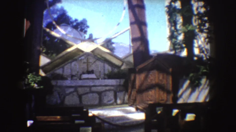 1962 California 8mm view of a rustic stone or stucco structure with a cross shape above the roofline, surrounded by trees under blue sky.