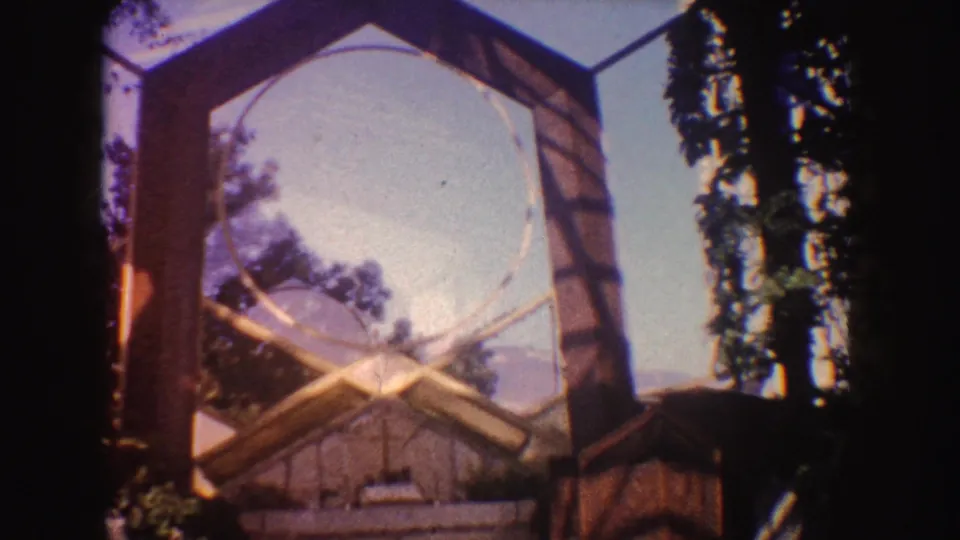 Closer 1962 California home-movie view of the chapel frame, stonework, trees, and rustic side structure.