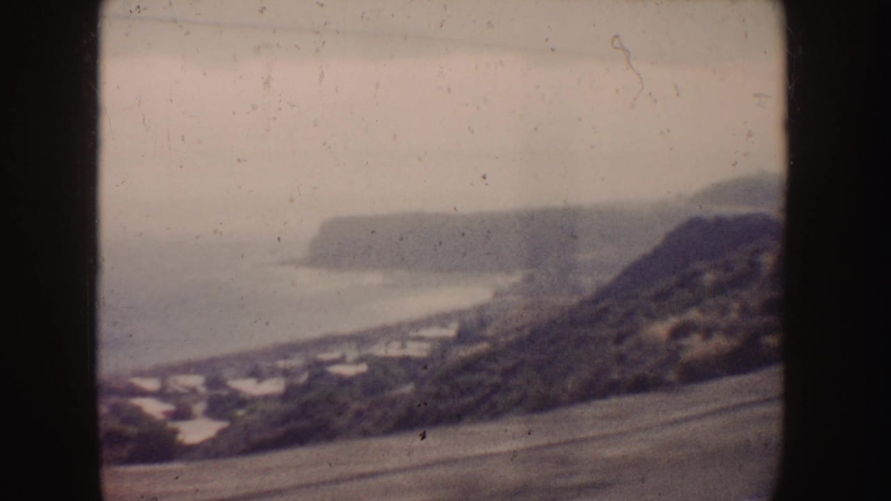 1962 8mm California coastal drive view with a hillside, road, shoreline, and distant cliffs seen through the vehicle window.