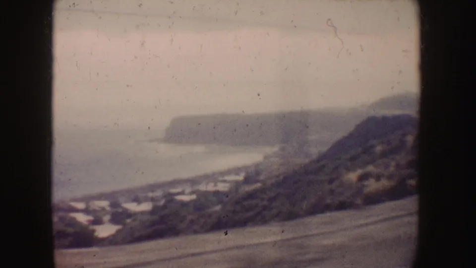 1962 8mm California coastal drive view with a hillside, road, shoreline, and distant cliffs seen through the vehicle window.