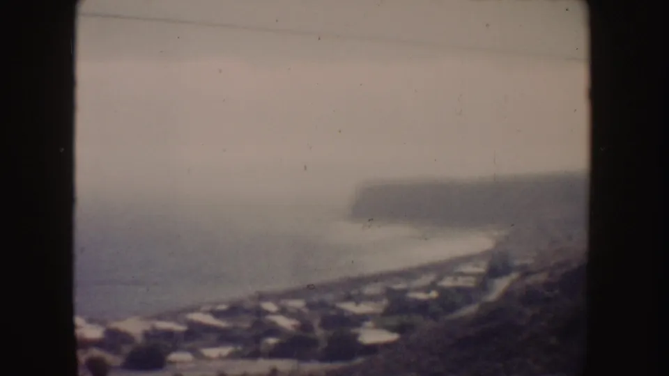 Soft 8mm view of the California coast, showing ocean, a shoreline road, hillside homes, and cliffs in the distance.