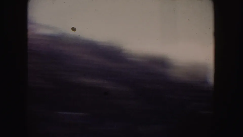 Motion-blurred 1962 8mm frame from a California coastal drive, with dark hillside foreground and pale ocean or sky beyond.