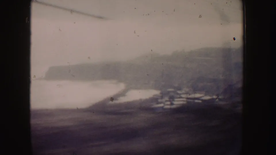 California coast seen from a moving car in 1962, with a bright shoreline, hillside houses, ocean water, and distant cliffs.