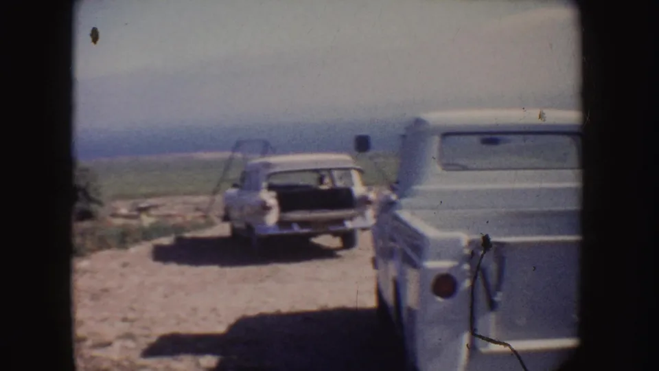 1962 California home-movie frame showing a station wagon and pickup truck parked along a rural dirt road near open fields.