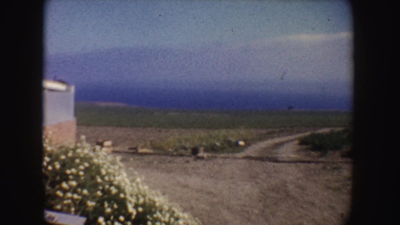 1962 California home-movie frame showing a dirt road or path near open fields, with a small roadside structure at left and hazy sky beyond.