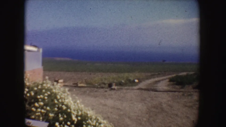 1962 California home-movie frame showing a dirt road or path near open fields, with a small roadside structure at left and hazy sky beyond.