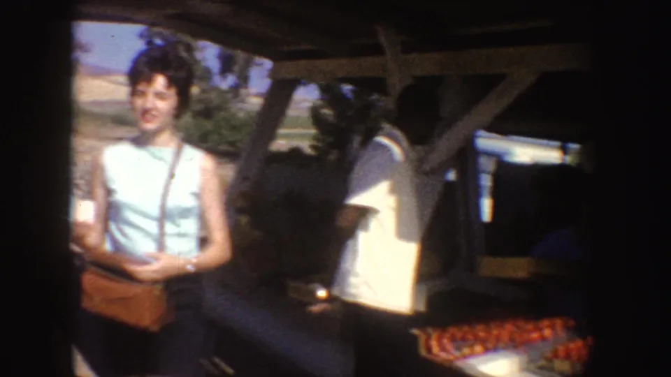 A woman in a light blue sleeveless top and dark shorts stands by a roadside fruit stand in California, with crates of oranges visible nearby.