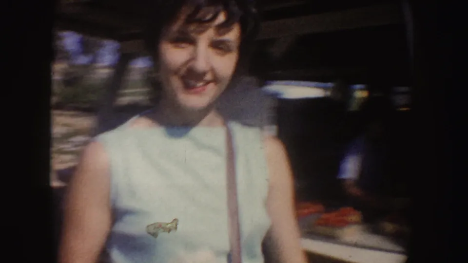 Closer 8mm frame of the smiling woman wearing a light blue sleeveless top and shoulder purse near the fruit stand display.