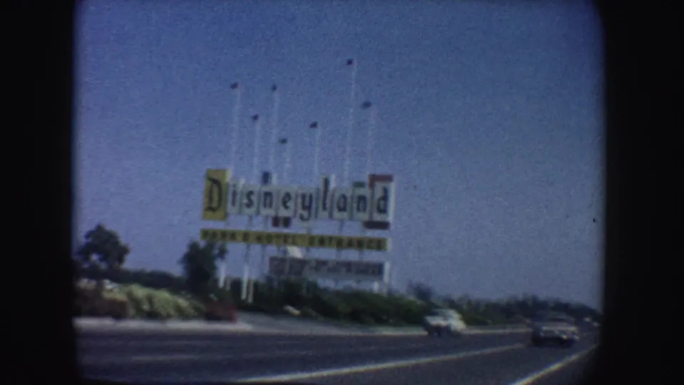 Highway view of the Disneyland sign in California, with cars on the road and flags above the sign against a bright blue sky.