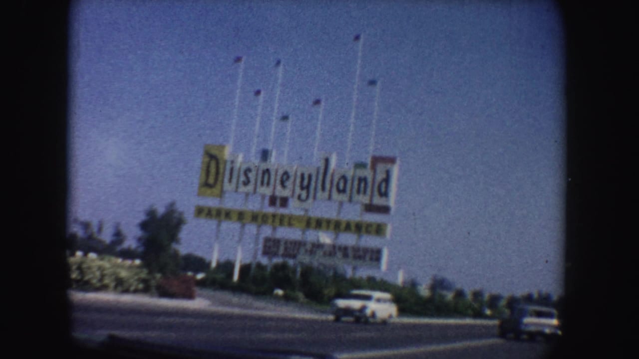 1962 California 8mm frame showing the Disneyland Park and Hotel Entrance sign, flagpoles, blue sky, landscaping, and passing cars.