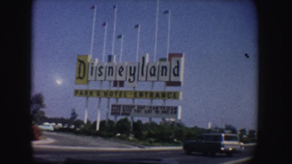 1962 California home-movie frame of the Disneyland Park and Hotel Entrance sign, roadside landscaping, flagpoles, and a car passing at right.