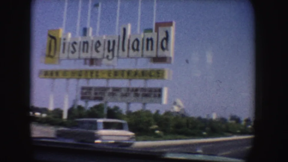 Closer 1962 California 8mm view of the Disneyland entrance sign, flagpoles, planted verge, and a mid-century car passing in front.