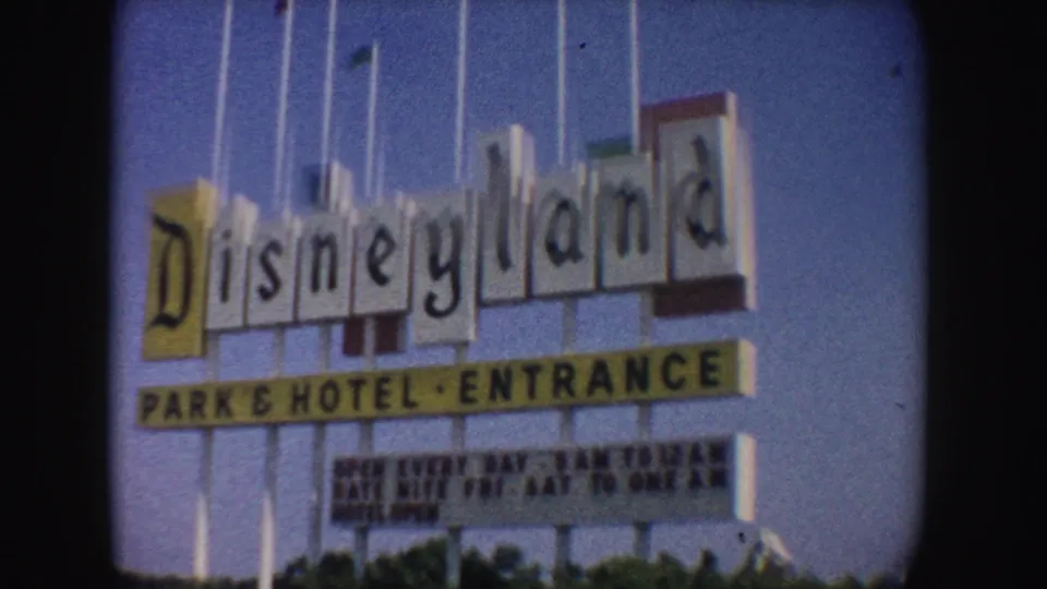 Close 1962 California 8mm frame of the Disneyland Park and Hotel Entrance sign with tall flagpoles, blue sky, and planted roadside below.