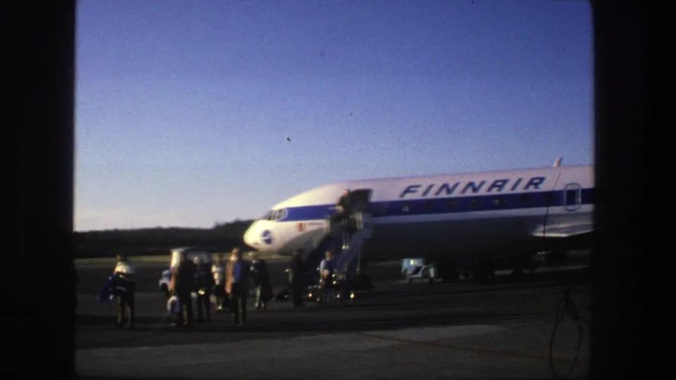 1975 Stockholm airport tarmac view with a Finnair aircraft, mobile stairs, passengers, crew, and a small service vehicle.