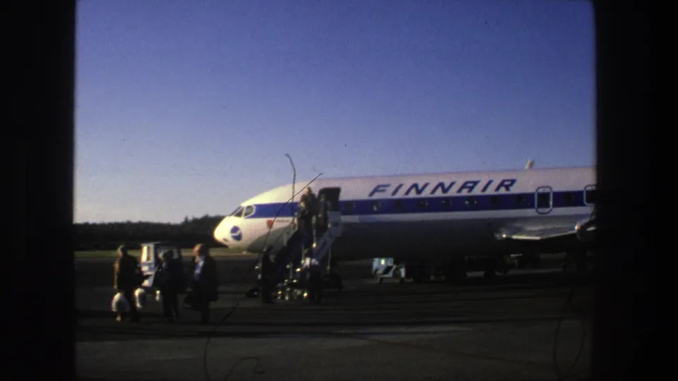 Passengers move across the Stockholm tarmac near a blue-and-white Finnair aircraft and mobile crew stairs in 1975 footage.