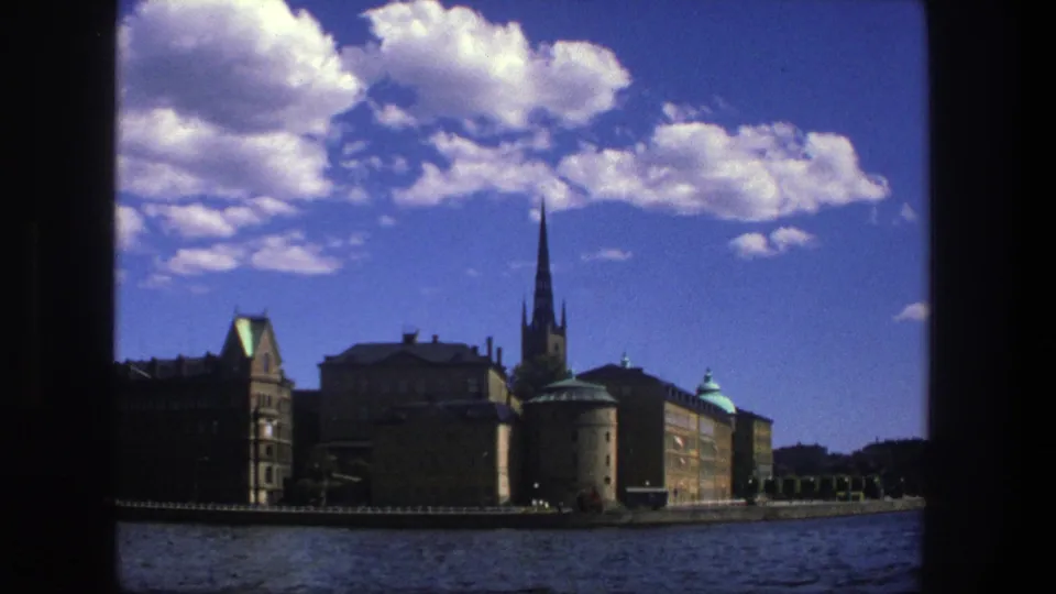 Stockholm waterfront view from 1975 metadata, with water, historic buildings, a prominent church spire, scattered clouds, and bright blue sky.
