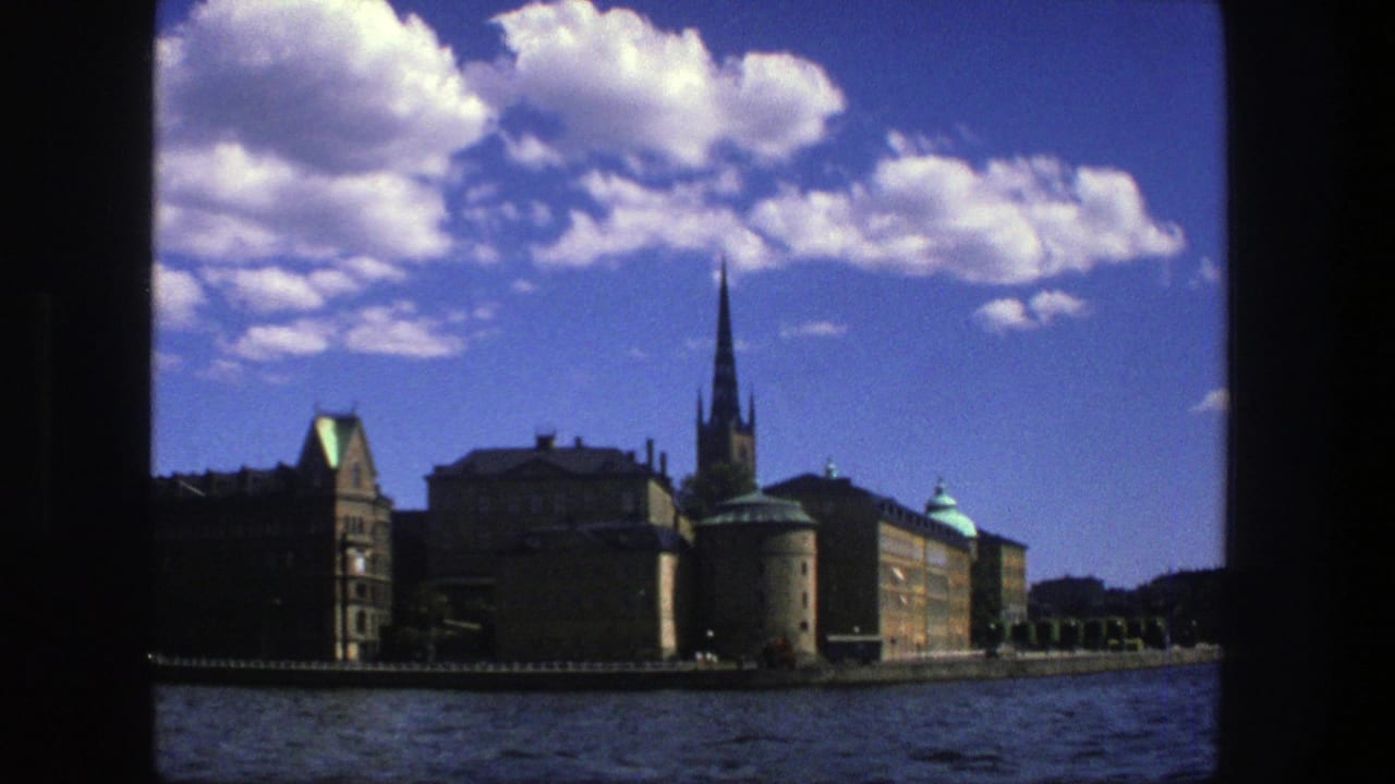 1975 8mm view of Stockholm waterfront architecture from the water, with a tall church spire, green roof details, and clouds in a deep blue sky.