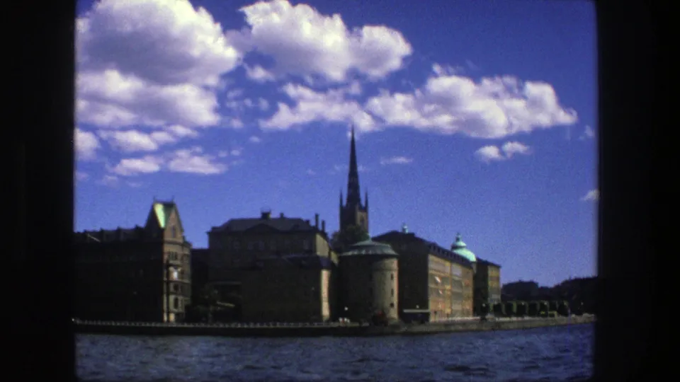 1975 8mm view of Stockholm waterfront architecture from the water, with a tall church spire, green roof details, and clouds in a deep blue sky.