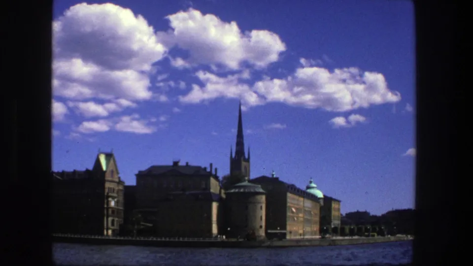 Stockholm waterfront scene on 8mm film, showing older masonry buildings, a narrow church spire, blue water, and a cloud-filled sky.