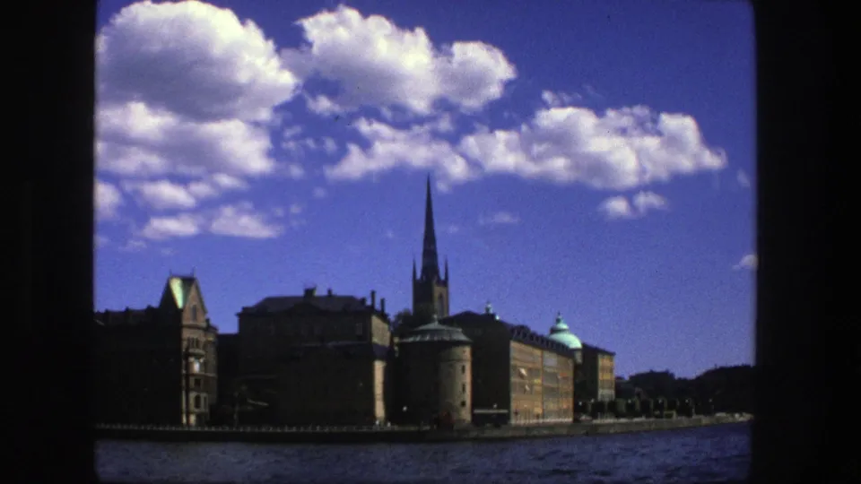 1975 Stockholm waterfront footage with buildings in partial shadow, a tall spire, rippling water, and bright clouds overhead.