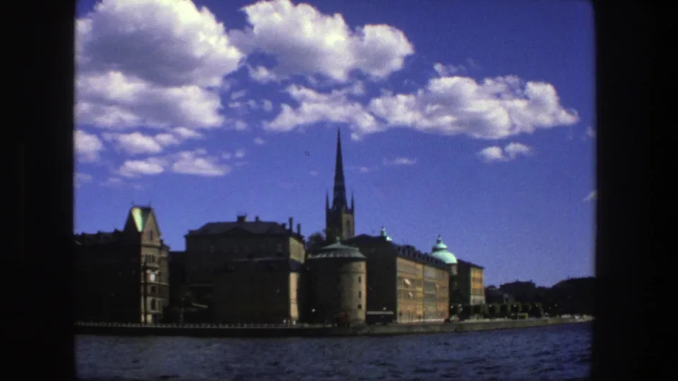 Stockholm waterfront view from 1975 8mm footage, with the church spire centered near older buildings and blue water in the foreground.