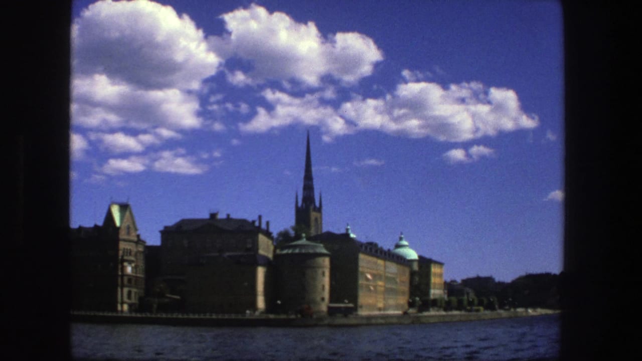 1975 Stockholm boat view showing waterfront buildings, a tall church spire, water, clouds, and dark 8mm frame borders.