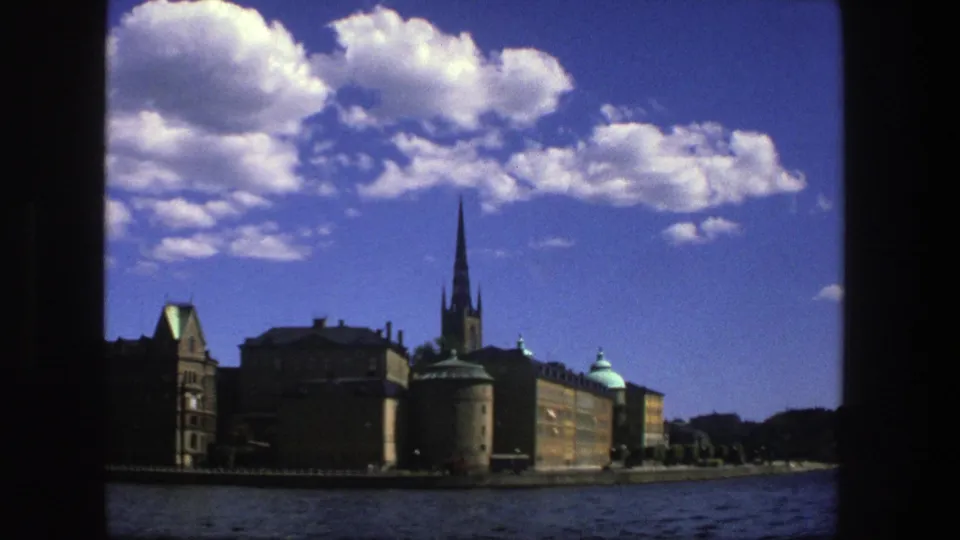 1975 Stockholm boat view showing waterfront buildings, a tall church spire, water, clouds, and dark 8mm frame borders.