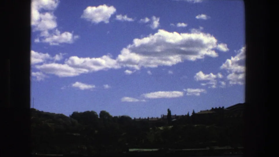 1975 Stockholm waterside view with wooded shoreline, low distant structures, and large white clouds filling the upper frame.