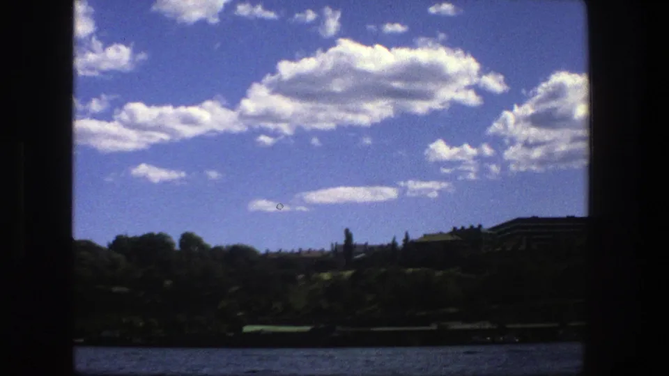 1975 Stockholm boat-view frame with water in the foreground, dark tree line, distant buildings, and bright clouds above.