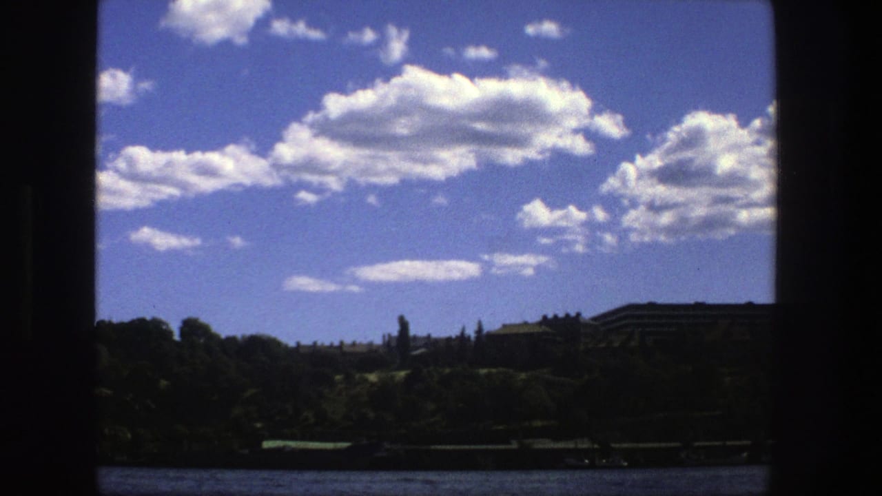 1975 Stockholm boat view with open water, a shadowed shoreline, distant buildings, and bright clouds behind dark window-like borders.