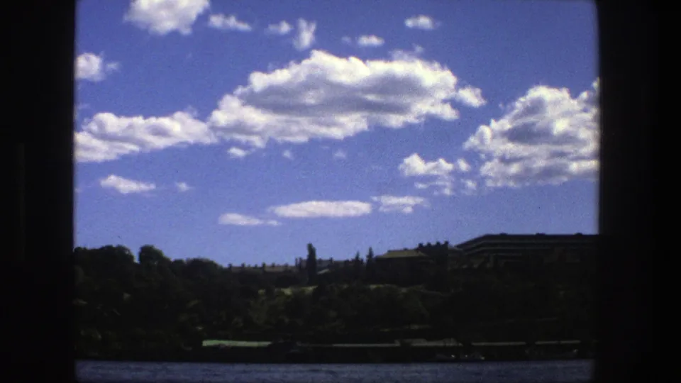 1975 Stockholm boat view with open water, a shadowed shoreline, distant buildings, and bright clouds behind dark window-like borders.
