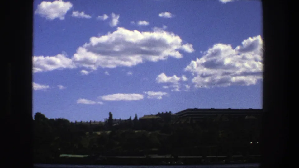 Stockholm shoreline from a boat, with calm water, trees, distant low buildings, and strong blue sky visible through dark borders.