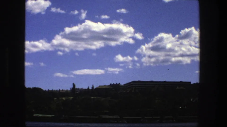 1975 Stockholm waterfront view showing shadowed trees, distant buildings, open water, and a saturated blue sky with clouds.