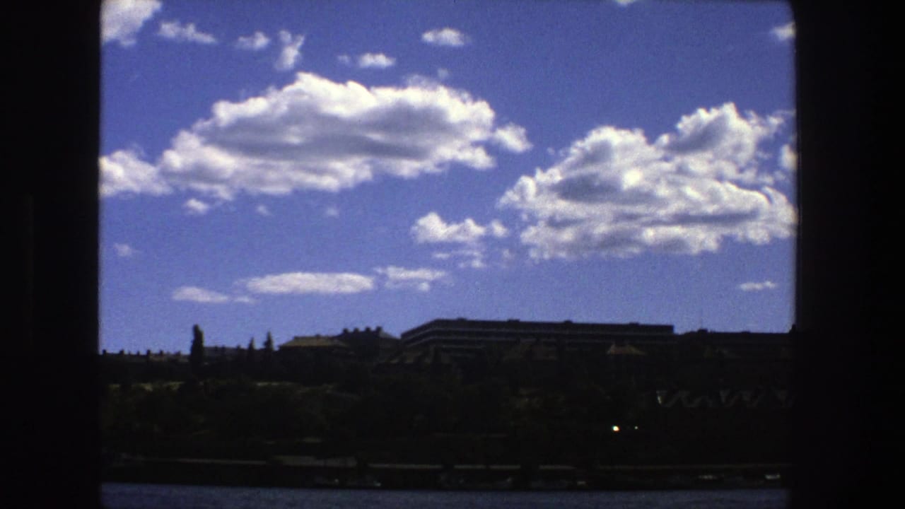 Cloud-filled blue sky above dark trees and low buildings, divided by a vertical black frame.