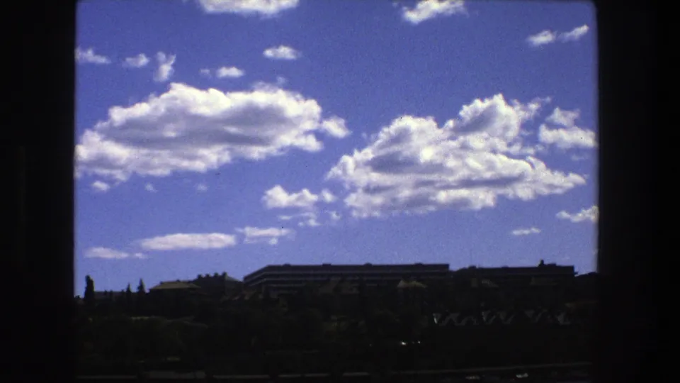 1975 Stockholm 8mm still with bright cumulus clouds over silhouetted trees and buildings, framed by dark edges.