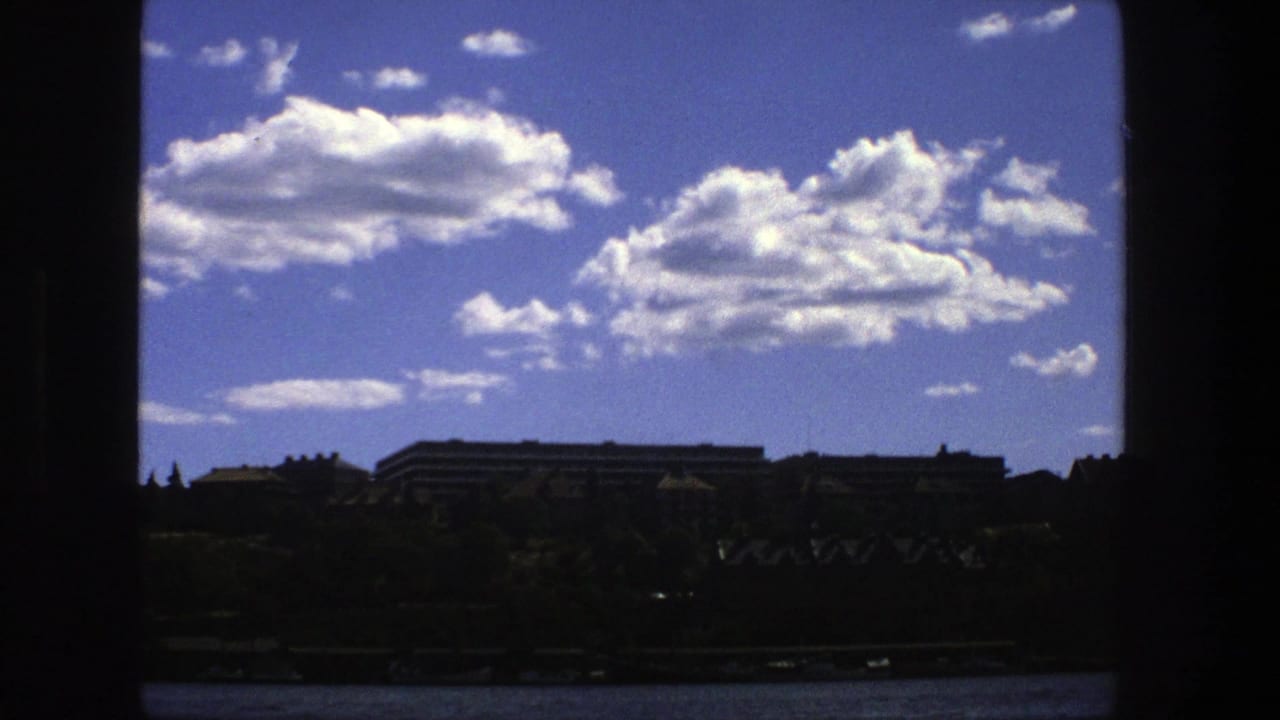 1975 Stockholm home-movie frame looking through a dark window toward clouds, distant buildings, trees, and a waterline.