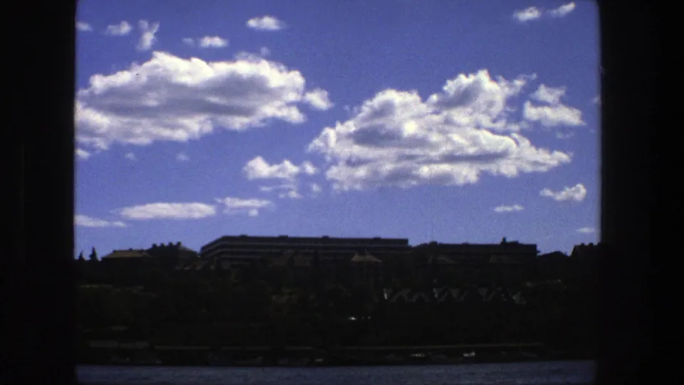 1975 Stockholm home-movie frame looking through a dark window toward clouds, distant buildings, trees, and a waterline.