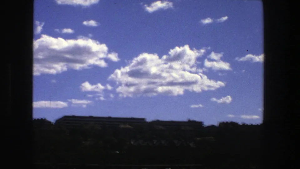 1975 Stockholm view through a dark window frame, with bright clouds over distant buildings, trees, and a thin strip of water.