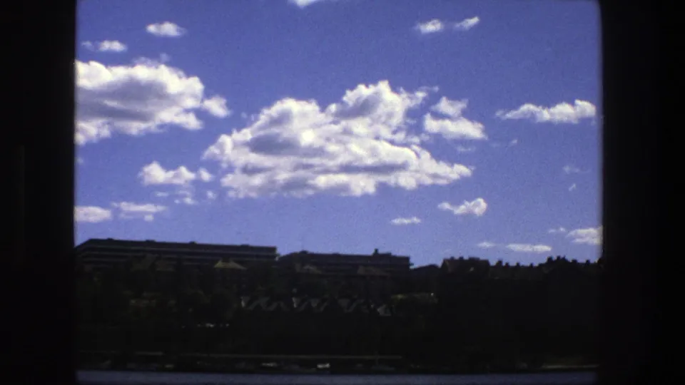 1975 Stockholm 8mm frame with a dark interior window edge, large clouds, distant urban buildings, trees, and water below.