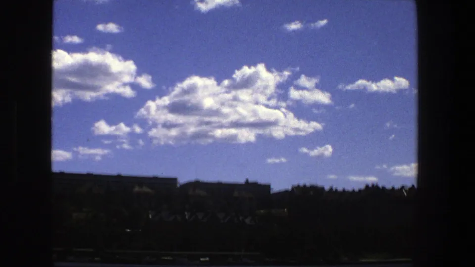 1975 Stockholm home-movie view through a silhouetted window, showing clouds over a distant waterfront and tree-lined buildings.