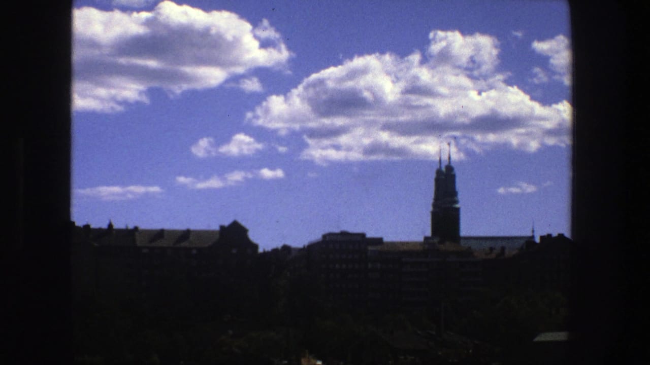 1975 8mm view of Stockholm with bright clouds over dark rooftops, trees, and a distant church tower framed by a window edge.