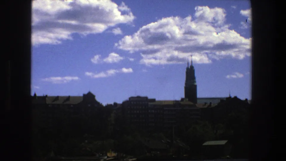 A 1975 Stockholm home-movie frame shows a dark window edge cutting into a skyline of rooftops, trees, clouds, and a distant tower.