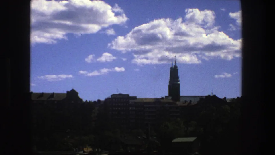 Late clip frame from the 1975 Stockholm 8mm view, with clouds above shadowed buildings, trees, and the tower near the right edge.