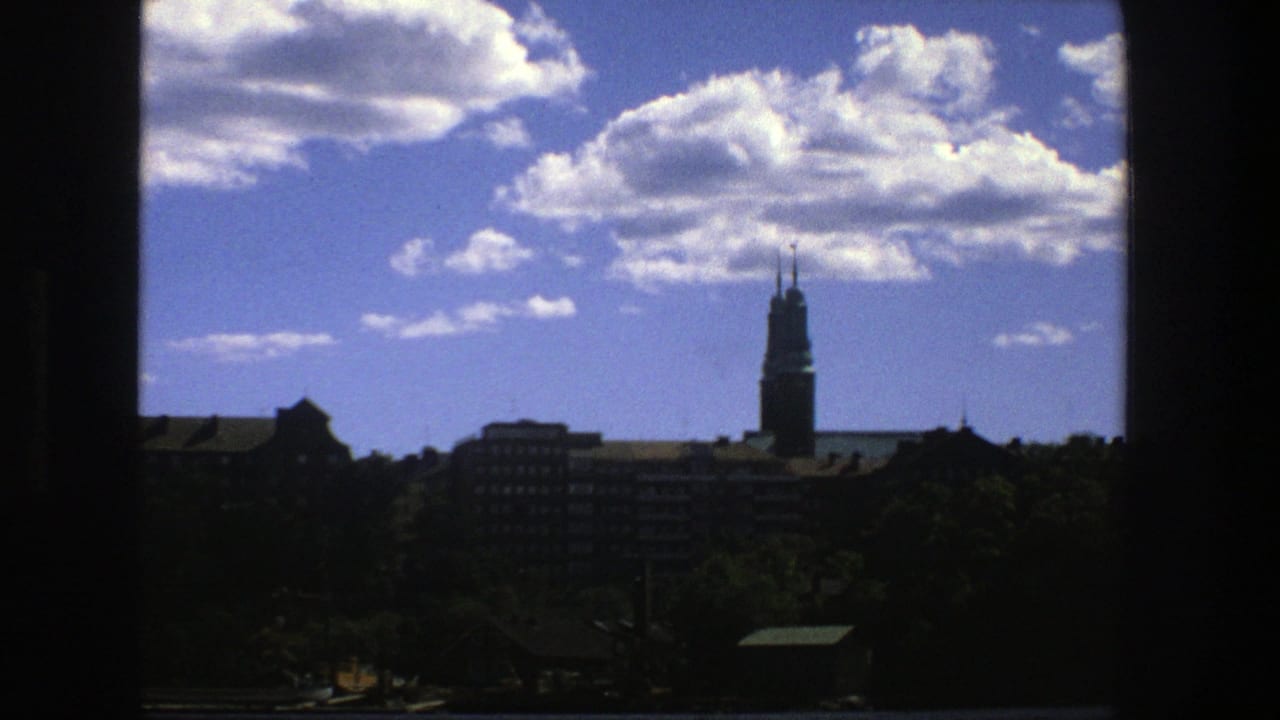 1975 8mm view of a Stockholm skyline with a tall church spire, waterfront trees, and cumulus clouds seen through dark framing.