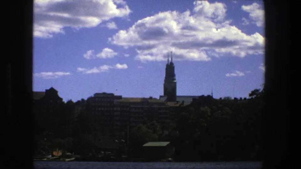 1975 Stockholm skyline view with a tall spire at right, mid-rise buildings, trees, and a bright cloud-filled sky.