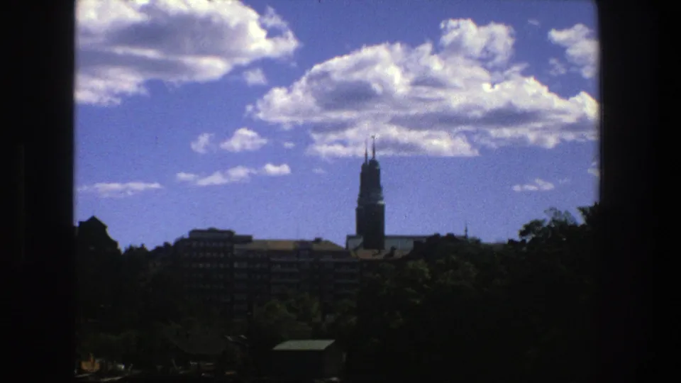 A 1975 8mm Stockholm view shows a prominent spire above buildings and trees, with dark framing around the bright sky.